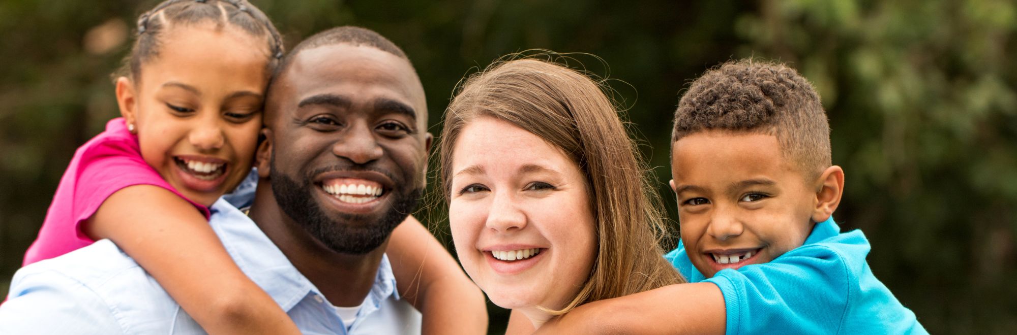 Smiling family of four outdoors, with two children hugging their parents.